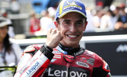 MotoGP rider in a red Lenovo racing suit, smiling with a raised fist, wearing a blue Michelin cap; cheering crowd in the background.