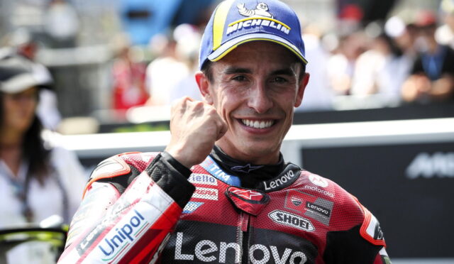 MotoGP rider in a red Lenovo racing suit, smiling with a raised fist, wearing a blue Michelin cap; cheering crowd in the background.