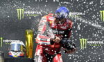 MotoGP rider in red Lenovo suit sprays champagne on a podium with Monster Energy banners in the backdrop, helmet in hand.