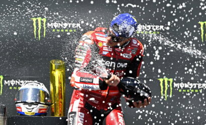 MotoGP rider in red Lenovo suit sprays champagne on a podium with Monster Energy banners in the backdrop, helmet in hand.