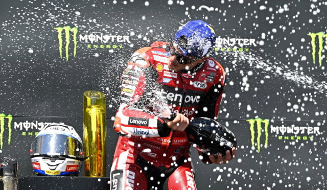 MotoGP rider in red Lenovo suit sprays champagne on a podium with Monster Energy banners in the backdrop, helmet in hand.