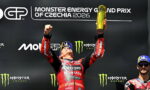 MotoGP rider on a podium, arms raised holding a gold trophy, in red racing suit, with a Monster Energy Czechia 2026 backdrop and a teammate on the right.