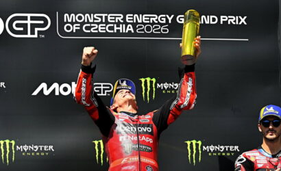 MotoGP rider on a podium, arms raised holding a gold trophy, in red racing suit, with a Monster Energy Czechia 2026 backdrop and a teammate on the right.