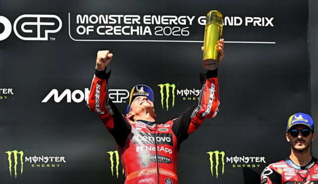 MotoGP rider on a podium, arms raised holding a gold trophy, in red racing suit, with a Monster Energy Czechia 2026 backdrop and a teammate on the right.