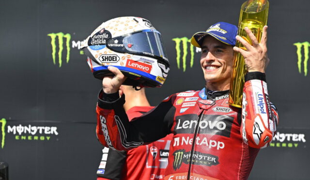Motorcyclist in red Lenovo racing suit holds a tall gold trophy and helmet, smiling on the podium with Monster Energy logos in the background.