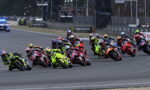 Group of motorcycle racers leaning into a turn on a race track, with colorful bikes and helmets and spectators in the background.
