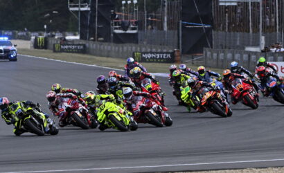 Group of motorcycle racers leaning into a turn on a race track, with colorful bikes and helmets and spectators in the background.
