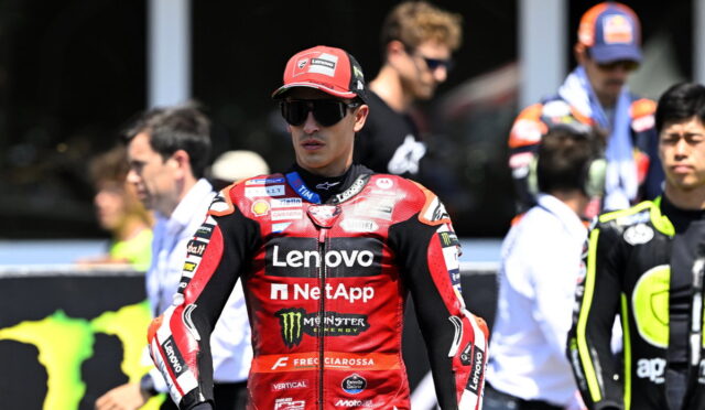 MotoGP rider in red Lenovo NetApp suit wearing black sunglasses and a red cap, standing in the pit area with crew in the background.