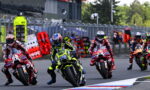 Motorcycle race: riders in colorful gear lean into a corner on a track, behind safety barriers and pit area.