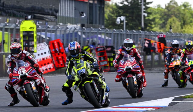 Motorcycle race: riders in colorful gear lean into a corner on a track, behind safety barriers and pit area.
