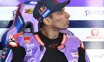 MotoGP rider in purple leather suit seated in a racing pit area, looking to the right; cap with Red Bull/Yamaha logos visible behind sponsor backdrop.