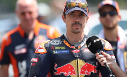 MotoGP rider in Red Bull racing suit being interviewed, wearing a cap and sunglasses, with a microphone held near his chest.