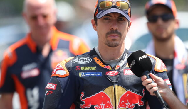 MotoGP rider in Red Bull racing suit being interviewed, wearing a cap and sunglasses, with a microphone held near his chest.