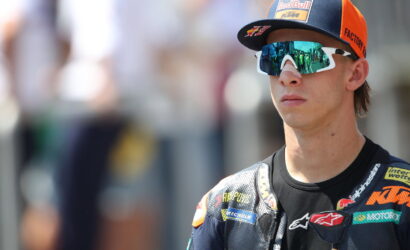 Young motorcycle racer in a sponsor-studded riding suit and orange Red Bull cap, wearing mirrored sunglasses.