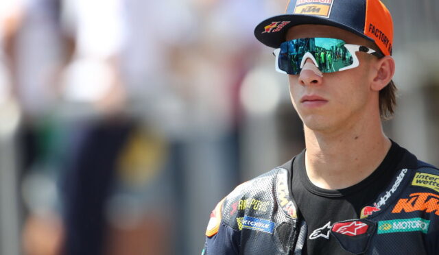 Male motorcycle racer wearing a Red Bull KTM cap and mirrored sunglasses, looking ahead in a racing suit with sponsor logos