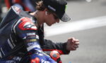 Motorcycle racer in a Yamaha racing suit kneeling and looking down, wearing a cap and wireless earbud in one ear.
