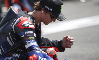 Motorcycle racer in a Yamaha racing suit kneeling and looking down, wearing a cap and wireless earbud in one ear.
