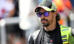 MotoGP rider wearing a neon green racing suit and Monster cap, looking pensively to the side with purple sunglasses.