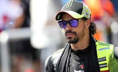 MotoGP rider wearing a neon green racing suit and Monster cap, looking pensively to the side with purple sunglasses.