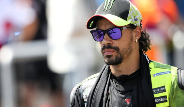 MotoGP rider wearing a neon green racing suit and Monster cap, looking pensively to the side with purple sunglasses.