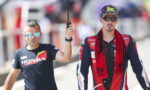 Two men walk side by side outdoors; a man in a Honda t-shirt holds an umbrella over a rider in a red-blue racing suit and cap.
