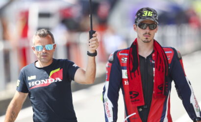 Two men walk side by side outdoors; a man in a Honda t-shirt holds an umbrella over a rider in a red-blue racing suit and cap.