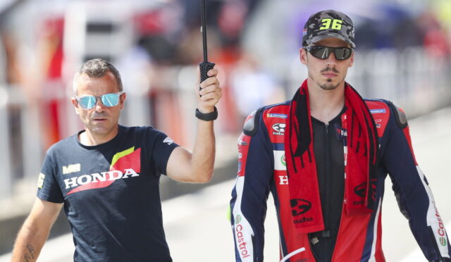 Two men walk side by side outdoors; a man in a Honda t-shirt holds an umbrella over a rider in a red-blue racing suit and cap.