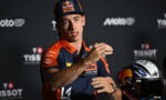 MotoGP rider in KTM Red Bull gear speaking at a press conference, hand gesturing, helmet on the table.