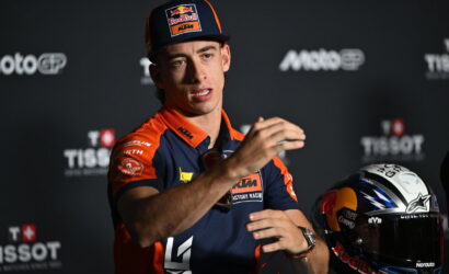 MotoGP rider in KTM gear speaks at a press event, gesturing with hands, helmet resting nearby on the tableLux 49