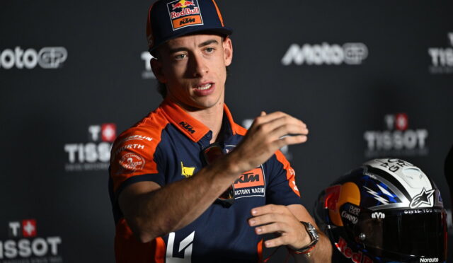MotoGP rider in KTM gear speaks at a press event, gesturing with hands, helmet resting nearby on the tableLux 49