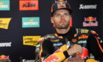 MotoGP rider in Red Bull KTM gear at a press conference, hands clasped, sponsor logos visible.