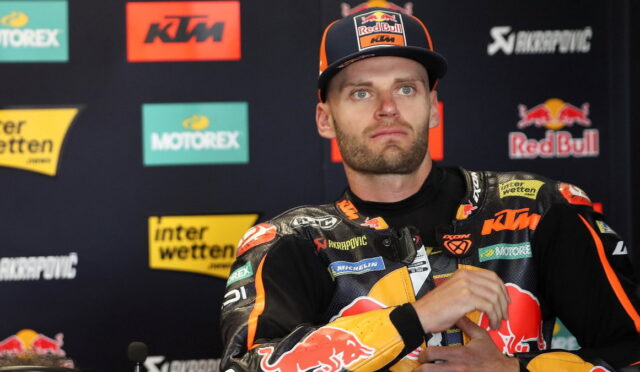 MotoGP rider in Red Bull KTM gear at a press conference, hands clasped, sponsor logos visible.