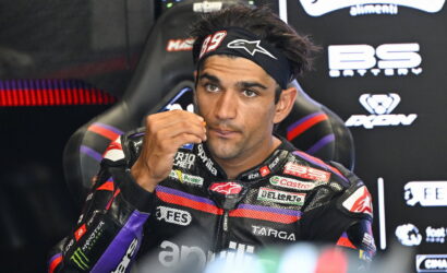 Moto racer wearing #29 headband sits in garage, adjusting his glove near his mouth, with sponsor logos in the background
