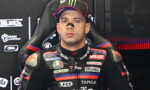 Moto racer in an Aprilia racing suit with sponsor patches, seated in a team chair and wearing a Monster cap.
