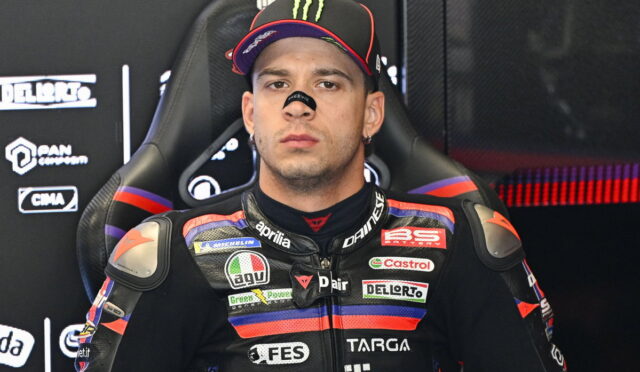 Moto racer in an Aprilia racing suit with sponsor patches, seated in a team chair and wearing a Monster cap.