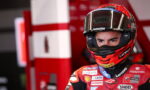MotoGP rider wearing red race suit and helmet, focused stare, in a pit area with red equipment behind him