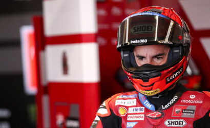 MotoGP rider wearing red race suit and helmet, focused stare, in a pit area with red equipment behind him