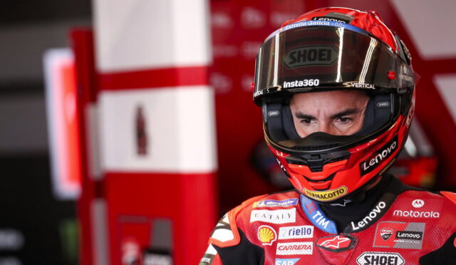 MotoGP rider wearing red race suit and helmet, focused stare, in a pit area with red equipment behind him