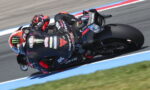 Moto racer leaning into a sharp turn on a black Aprilia bike, helmet featuring Monster Energy logo, on a racetrack with blue edging.