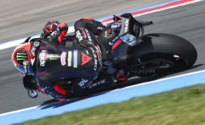 Moto racer leaning into a sharp turn on a black Aprilia bike, helmet featuring Monster Energy logo, on a racetrack with blue edging.