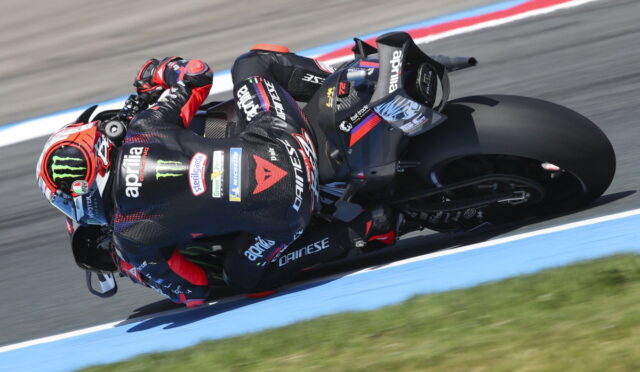 Moto racer leaning into a sharp turn on a black Aprilia bike, helmet featuring Monster Energy logo, on a racetrack with blue edging.