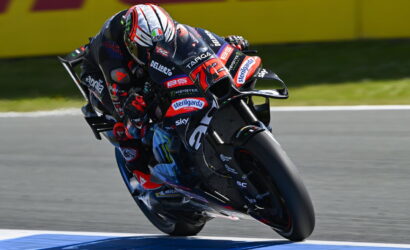 Racing motorcycle leaned into a turn at high speed, rider in full suit with sponsor logos on a track edge barrier behind.