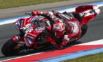 Red-suited motorcycle racer leans into a sharp turn on a sponsor-laden red race bike on a track edge.