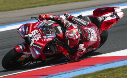 Red-suited motorcycle racer leans into a sharp turn on a sponsor-laden red race bike on a track edge.
