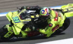 Motorcycle racer in neon yellow suit leaning into a sharp left turn on a bright yellow Ducati race bike, marked with sponsor logos, on a track with blue and white curbs.
