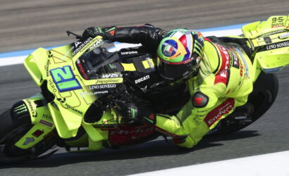 Motorcycle racer in neon yellow suit leaning into a sharp left turn on a bright yellow Ducati race bike, marked with sponsor logos, on a track with blue and white curbs.