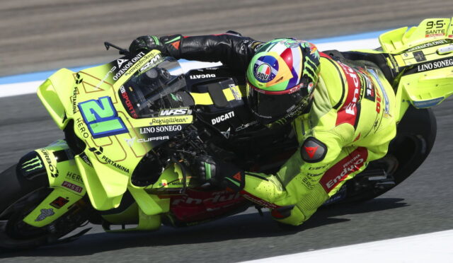 Motorcycle racer in neon yellow suit leaning into a sharp left turn on a bright yellow Ducati race bike, marked with sponsor logos, on a track with blue and white curbs.