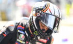 Racer in a black suit with sponsor patches, wearing a colorful full-face helmet and visor down.