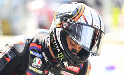 Racer in a black suit with sponsor patches, wearing a colorful full-face helmet and visor down.