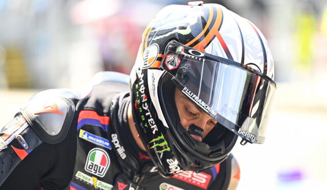 Racer in a black suit with sponsor patches, wearing a colorful full-face helmet and visor down.
